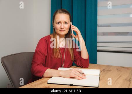 Une jeune femme aux longs cheveux blonds et un pull-over rouge écrit dans un livre tout en parlant sur son téléphone cellulaire et assis à une table en bois. Banque D'Images