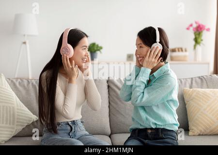 Une jeune femme asiatique heureuse et sa mère mature dans un casque qui écoutent de la musique ensemble, assis sur un canapé à la maison Banque D'Images