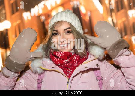 portrait d'une charmante femme dans une rue enneigée, portant une veste d'hiver, souriant avec joie contre les lumières de la ville, elle regarde le Banque D'Images