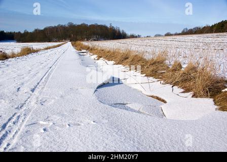 Route de terre avec des feuilles de glace fissurées entre les terres arables dans un paysage rural suédois avec de la neige au début du printemps. Banque D'Images