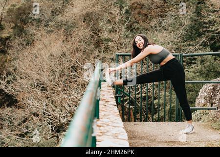 Jeune femme sur des vêtements de sport qui s'étire la jambe sur un trottoir au milieu d'une forêt automnale tout en souriant à l'appareil photo, à l'espace de copie, au centre de fitness Banque D'Images