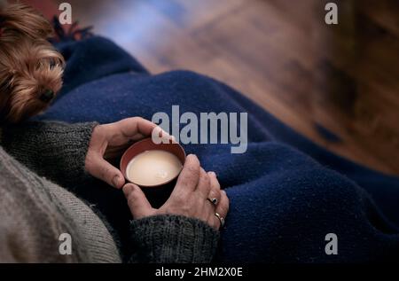 Une femme assise entend un animal couvrant ses jambes avec une couverture bleue et tient une tasse de thé chaud avec du lait dans ses mains. Banque D'Images