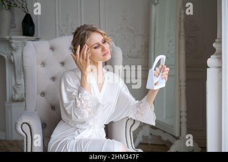 Tête magnifique femme blonde d'âge moyen, assise dans un fauteuil dans la salle de séjour, touchant son visage, regardant le reflet dans le miroir, faisant la beauté du matin.Concept de soins de la peau à la maison Banque D'Images