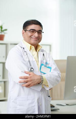 Portrait d'un médecin mature positif en blouse de travail blanche croisant les bras et souriant à la caméra Banque D'Images