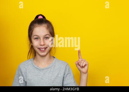 Portrait en gros plan d'une fille dans un t-shirt gris pointant vers le haut avec son doigt, isolé sur un fond jaune vif. Prise de décision et éducation des adolescents Banque D'Images