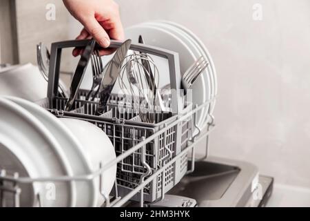 Gros plan de la main féminine tenant le panier à couverts complet avec un couteau propre, une fourchette et un fouet.Chargement, vidage ou déchargement d'une machine à lave-vaisselle automatique ouverte avec des ustensiles propres dans la cuisine maison Banque D'Images