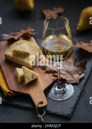 Un verre de vin blanc a été servi avec du fromage dans une planche à découper sur fond sombre. Pique-nique d'automne avec fromage, vin et feuilles sèches dans un style rustique. Banque D'Images