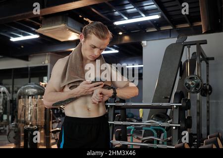 Athlète musclé sérieux vérifiant les résultats sportifs sur l'application smartphone et la montre intelligente après une bonne séance d'entraînement dans la salle de fitness Banque D'Images