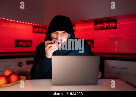 un hacker dans un sweat à capuche noir avec une capuche sur sa tête est assis à un ordinateur portable piquant une pomme rouge Banque D'Images