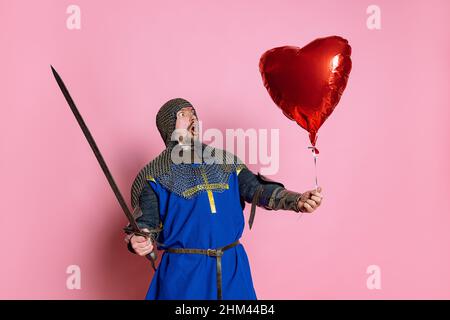 Portrait d'un homme choqué et excité, d'un guerrier médiéval ou d'un chevalier avec ballon rouge en forme de coeur isolé sur fond rose Banque D'Images