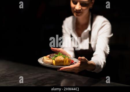 Le chef prépare une assiette avec repas au poste de commande dans la cuisine commerciale sur fond noir. Banque D'Images