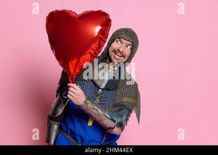 Portrait en demi-longueur d'un homme heureux et excité, guerrier médiéval en couverture protectrice avec ballon en forme de coeur rouge isolé sur fond rose Banque D'Images