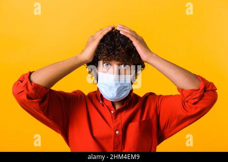 Oh mon Dieu.Portrait d'un jeune homme choqué dans un style décontracté avec masque médical chirurgical debout, tenant la tête et regardant l'appareil photo avec un visage incroyable.Prise de vue en studio, isolée sur l'orange Banque D'Images