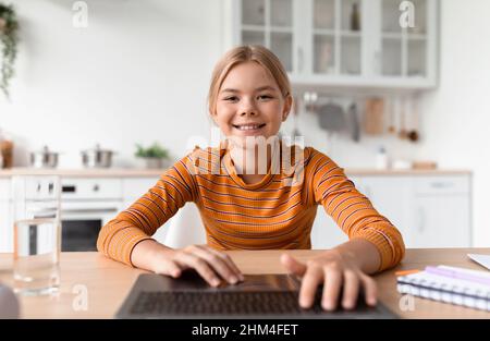 Bonne adolescente européenne blonde dactylographiant sur le clavier par ordinateur portable dans la cuisine intérieure confortable Banque D'Images