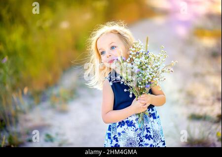 jolie petite fille mignonne avec des cheveux blonds marche à l'extérieur au coucher du soleil, tient un bouquet de fleurs sauvages dans ses mains, enfance heureuse Banque D'Images