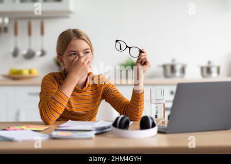 Une jeune fille blonde européenne fatiguée enlève ses lunettes et frotte les yeux à la table avec un ordinateur portable à l'intérieur de la cuisine Banque D'Images