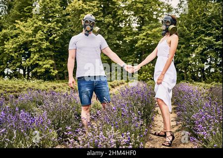 Portrait d'un jeune couple debout dans un champ de lavande portant des masques à gaz, tenant les mains.Personnes souffrant d'allergies et de fleurs de lavande.Concept d'allergie saisonnière Banque D'Images