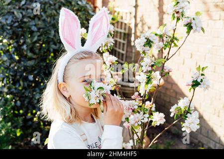 Adorable petite fille blonde dans les oreilles de lapin costume renifler la branche d'arbre en fleur à l'extérieur le jour du printemps. Enfant s'amusant sur la chasse aux œufs de Pâques dans le guar Banque D'Images