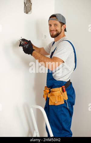 Joyeux électricien souriant à la caméra, tenant des pinces tout en travaillant sur le câblage dans une nouvelle maison Banque D'Images