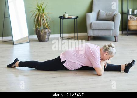 Flexible senior femme en noir leggins et beige t-shirt assis sur la ficelle sur le sol de la salle de séjour avec ses jambes divisées et étirées Banque D'Images