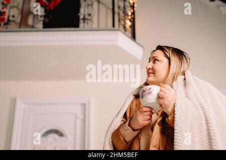 Vue latérale photo d'une femme dehors en vacances d'hiver regardant de côté se tenant près de la maison décorée avec des guirlandes gardant une tasse de boisson chaude. Banque D'Images