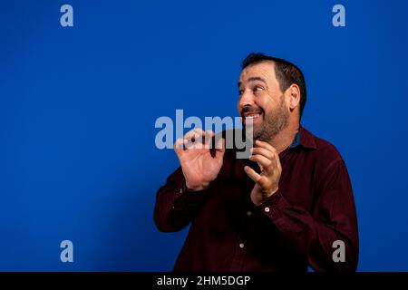 Wow et l'expression du visage choqué de l'homme timide barbu latin pendu de côté avec les mains près du visage se sentant embarrassé, isolé sur le studio bleu Banque D'Images
