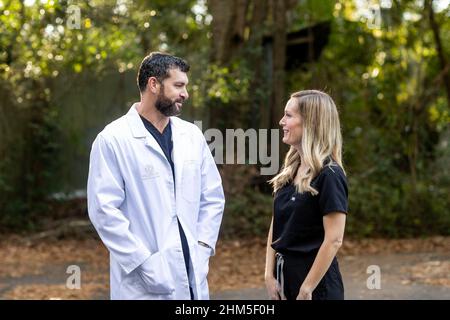 Un médecin aux cheveux foncés et une barbe en gommage noir et un manteau de laboratoire blanc debout à l'extérieur parlant à une infirmière praticienne médicale professionnelle Banque D'Images