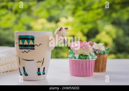 Magnifique tasse tendance en forme de lama avec boisson chaude et deux petits gâteaux aux fleurs de beurre sur table en bois blanc avec fond vert vif Banque D'Images