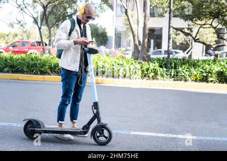 Latin homme avec des lunettes prenant un scooter avec son téléphone cellulaire et des écouteurs jaunes dans une rue de ville Banque D'Images