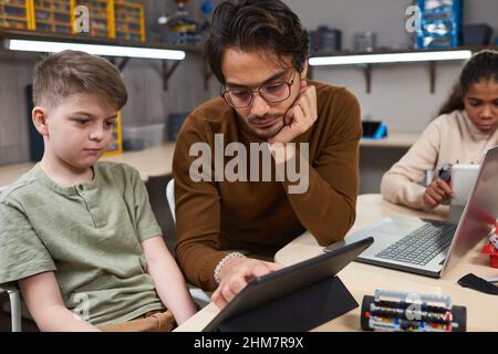 Portrait d'un jeune homme utilisant une tablette numérique tout en enseignant la robotique à divers groupes d'enfants dans l'école moderne Banque D'Images