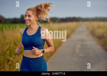 Posez une jeune femme en bonne santé en train de courir le long d'une route de campagne en passant la caméra avec un sourire heureux plein de vitalité dans un concept de style de vie actif Banque D'Images