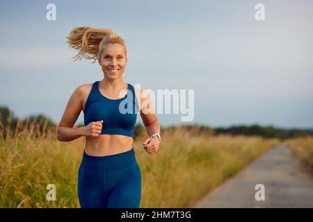 Posez une jeune femme en bonne santé en train de courir le long d'une route de campagne en passant la caméra avec un sourire heureux plein de vitalité dans un concept de style de vie actif Banque D'Images