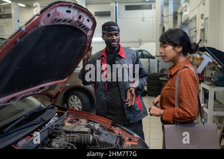 Portrait d'un mécanicien de voiture noir parlant à une cliente en se tenant debout près du capot ouvert dans un atelier de réparation automobile, espace de copie Banque D'Images