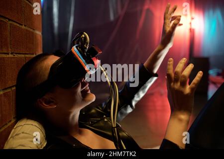 Gros plan d'une jeune femme qui s'étire les bras et sourit tout en jouant à un jeu en ligne dans des lunettes de réalité virtuelle dans une pièce sombre Banque D'Images