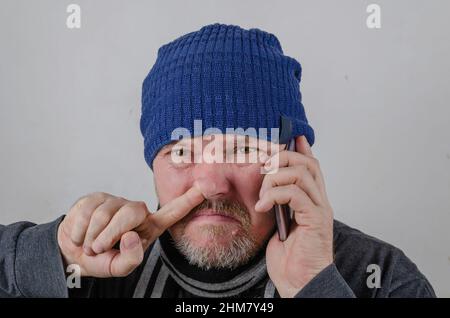 Un adulte qui se prend le nez en parlant au téléphone. Un homme d'âge moyen dans un chapeau bleu contre un mur de couleur claire. Banque D'Images