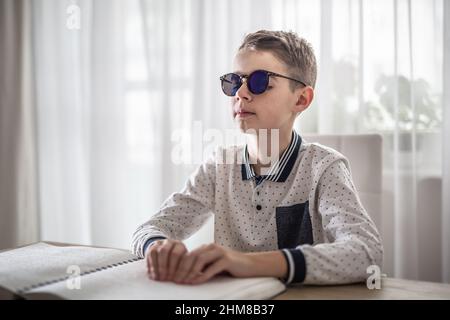 Un garçon aveugle en lunettes sombres lit un livre en braille. Banque D'Images