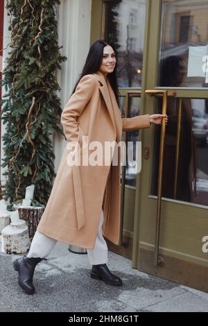 Jeune femme brunette portant un manteau beige ouvrant la porte en verre vert, entrant dans le café. Mode Street style Banque D'Images