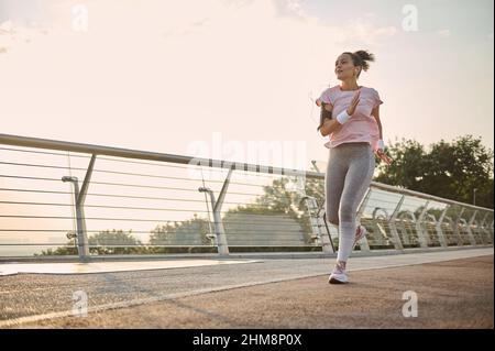 Longueur totale. Jeune femme sportive, jogger, coureur pratiquant le sport en plein air, courir rapidement le long du pont au-dessus du ciel de lever du soleil et de fond de rivière. Banque D'Images