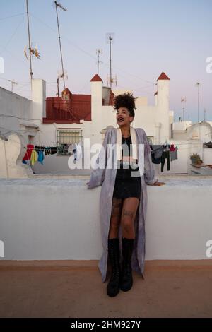 Femme afro-américaine dans des vêtements élégants riant et s'appuyant sur la barrière contre les maisons tout en se relaxant sur la terrasse le soir, Cadix, Espagne Banque D'Images