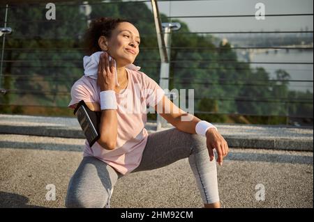 Belle athlète féminine ravie essuyant son cou de la sueur avec une serviette en éponge blanche, assis sur la position de squat sur le pont de la ville, se détendre après le jogging Banque D'Images
