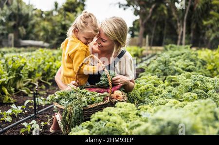 Collage dans le jardin. Bonne jeune mère souriant gaiement tout en portant sa fille et en cueillant du kale frais dans un jardin potager. Autosuffisant Banque D'Images