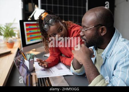 Femme d'affaires africaine discutant de documents avec un homme d'affaires lorsqu'ils sont assis à la table avec un ordinateur portable au bureau Banque D'Images