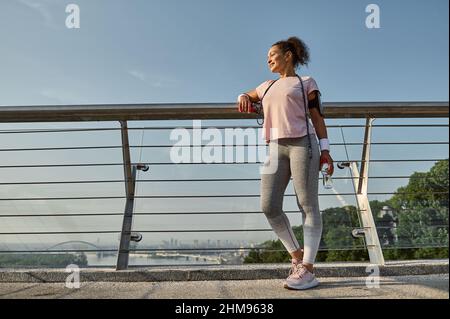 Portrait complet d'une sportswoman, athlète féminine avec corde à sauter, debout sur le pont moderne de la ville de verre, se détendre après l'entraînement cardio ou Banque D'Images