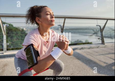 Déterminée sportive confiante femme avec un porte-smartphone sur son bras exécutant un entraînement de poids corporel, en utilisant la résistance élastique fitn Banque D'Images