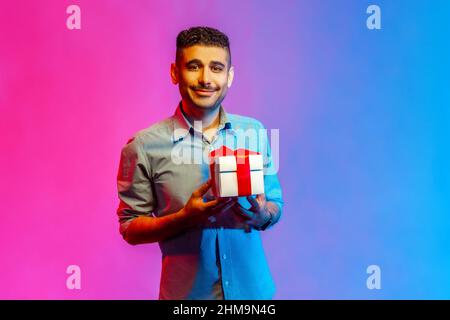 Portrait d'un homme souriant et souriant dans une chemise tenant un cadeau et regardant un appareil photo avec un sourire crasseux, des cadeaux et des bonus, la célébration. Studio d'intérieur isolé sur fond de néon coloré. Banque D'Images