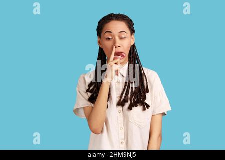 Portrait de femme impolie drôle avec des dreadlocks noirs rendant le visage fou avec la langue dehors et de choisir son nez, mauvaises manières, portant chemise blanche. Studio d'intérieur isolé sur fond bleu. Banque D'Images