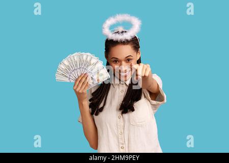 Positive angélique femme avec des dreadlocks noirs et halo au-dessus de la tête tenant le ventilateur de billets de dollars et pointant le doigt vers la caméra, choisissant le gagnant de loterie. Studio d'intérieur isolé sur fond bleu. Banque D'Images