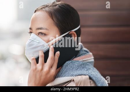Il est important de rester connecté pendant les périodes difficiles. Photo d'une jeune femme portant un masque tout en parlant au téléphone sur le balcon à la maison. Banque D'Images