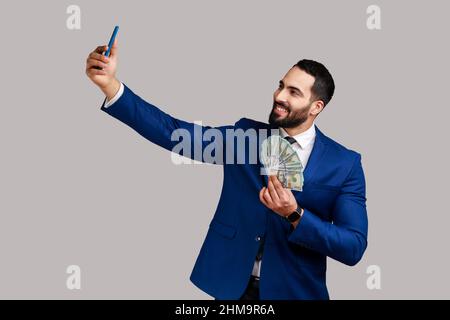 Homme barbu faisant le selfie avec de l'argent, se cachant face derrière un fan de billets de cent dollars, se vantant avec des revenus, faisant des cadeaux, portant le costume officiel. Prise de vue en studio isolée sur fond gris Banque D'Images