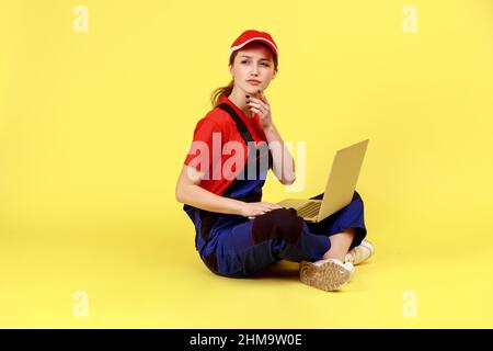 Portrait complet d'une travailleuse pensive assise sur le sol avec des jambes croisées et travaillant sur un ordinateur portable, regardant de côté, pensant, portant une combinaison. Studio d'intérieur isolé sur fond jaune. Banque D'Images